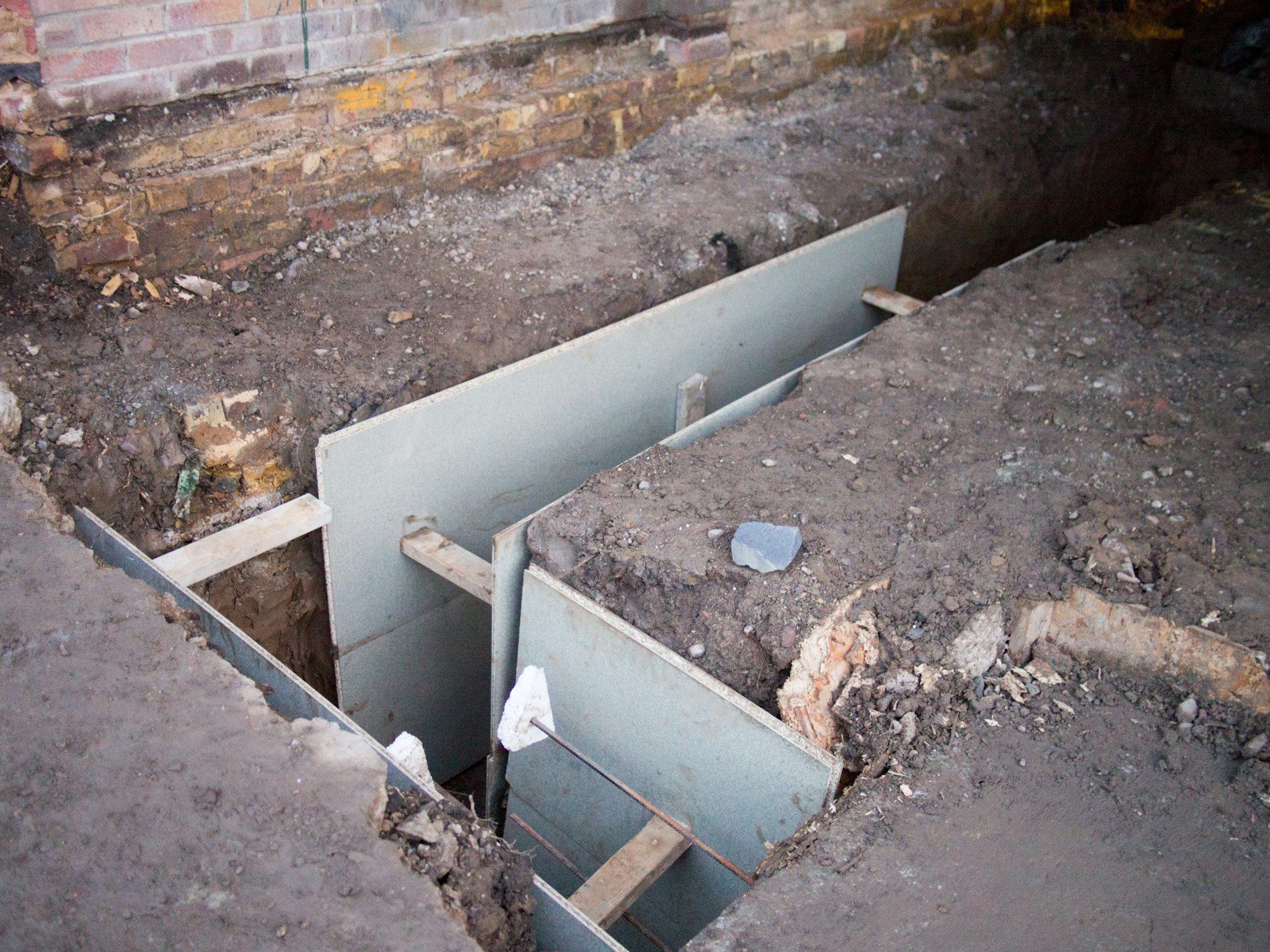 An open excavation pit beside a brick house wall with metal shuttering panels and soil.