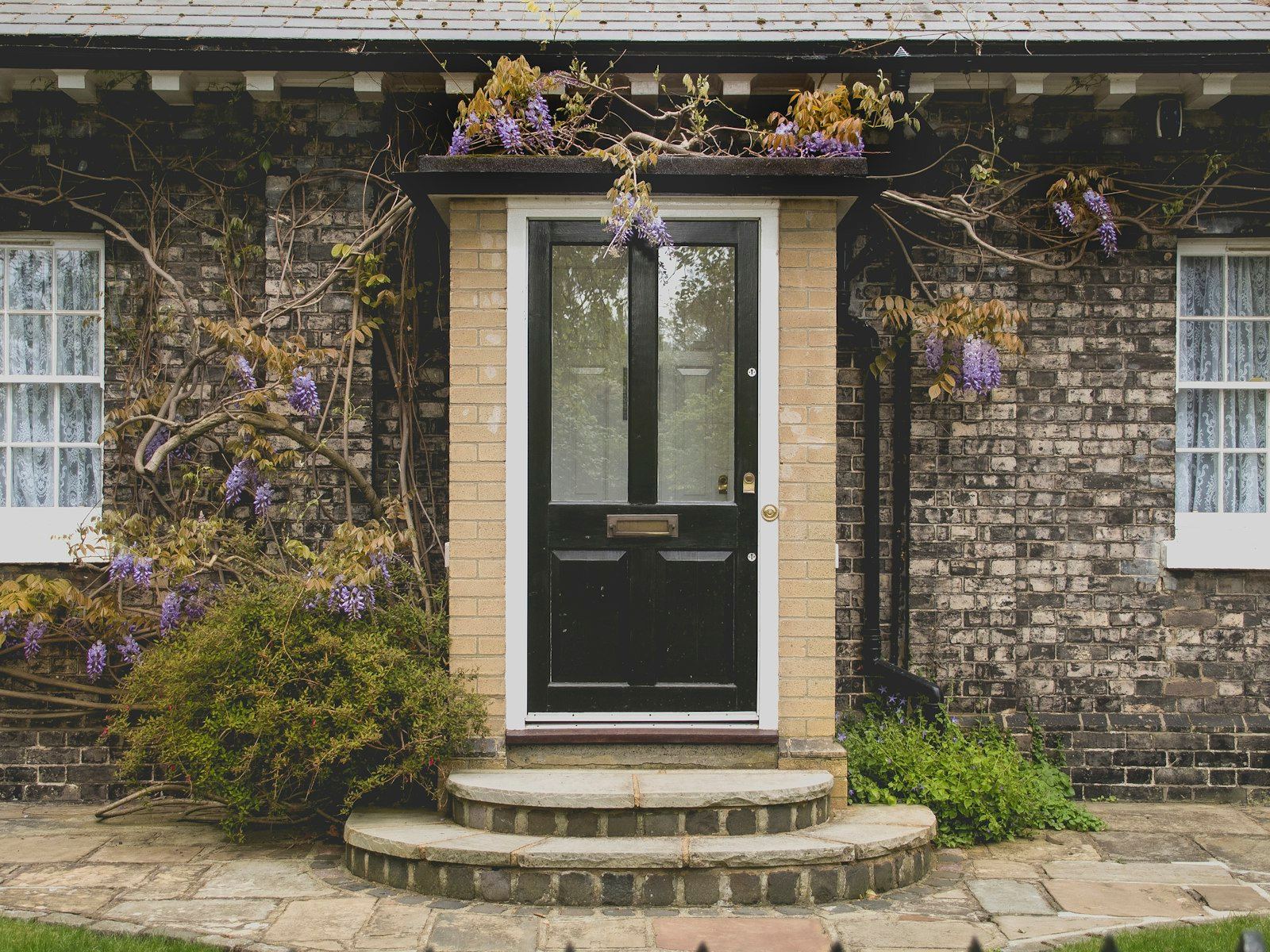 A black front door with tiled steps surrounded by wisteria on a brick cottage.