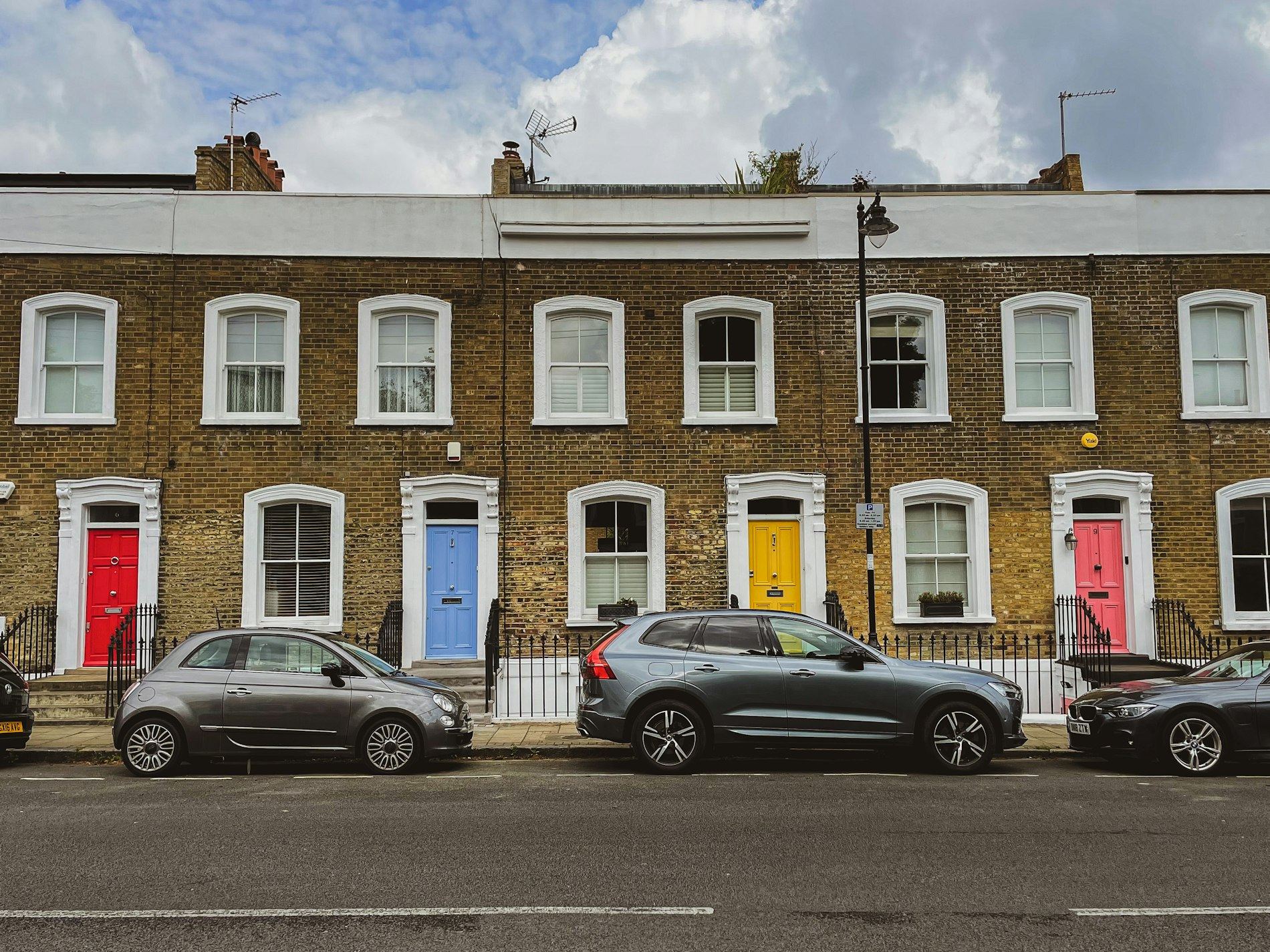 A row of two-storey brick terraced houses with colourful front doors and parked cars.